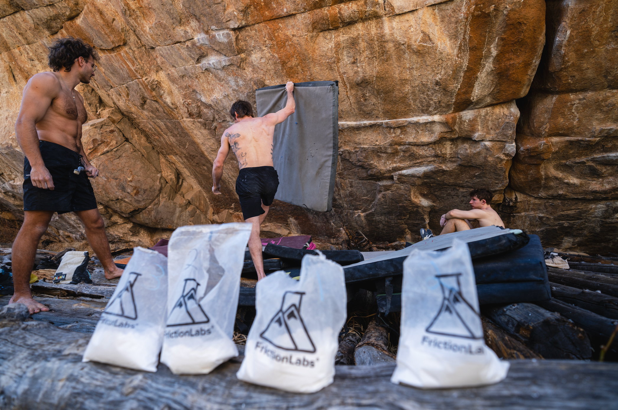 Three men are seen bouldering in front of a large rock face. Several white FrictionLabs climbing chalk bags are lined up in the foreground on the ground.